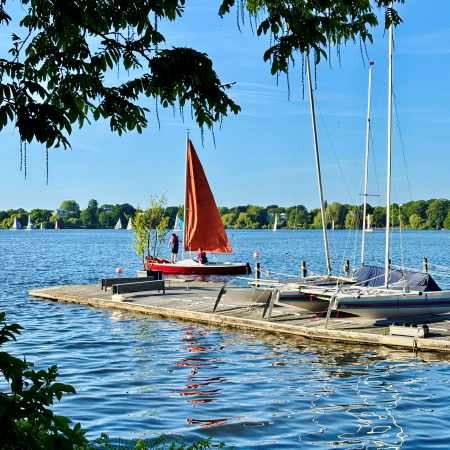 Ein Segelboot mit rotem Segel, das gerade von einem Steg an der Außenalster losfährt