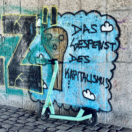 Graffiti an einer Wand in Planten un Blomen: Das Gespenst des Kapitalismus. Davor ein E-Scooter aus dem Leih-Bestand.