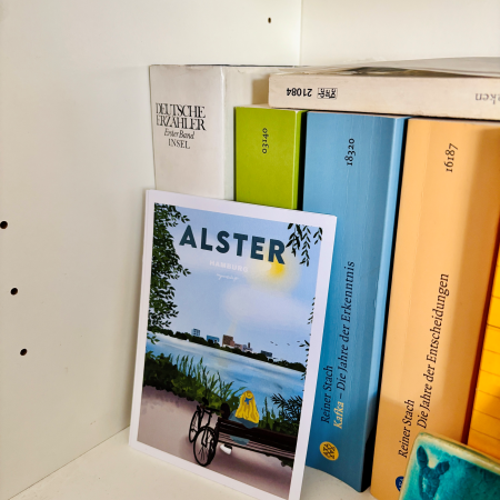Detail eines Buchregals mit einer Postkarte, auf der die Alster abgebildet ist und dahinter Bänden einer Kafka-Biografie