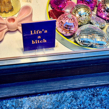 Ein Schild in einem Schaufenster, neben glitzernder Deko: Life's a bitch