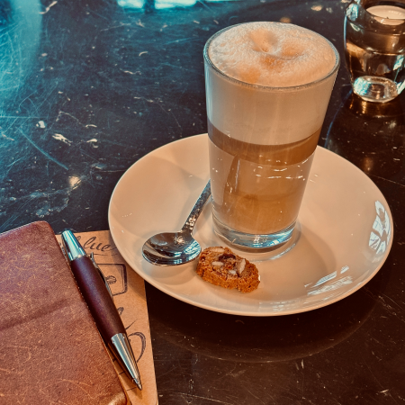 Ein Latte Macchiato, daben ein Notizbuch mit Kugelschreiber