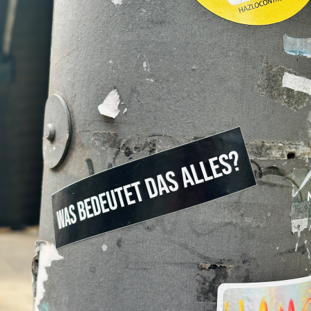 Ein Sticker an einem Laternenmast mit der Aufschrift: Was bedeutet das alles?